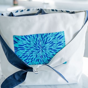 Puede incluir: Bolsa de lona blanca con un bolsillo de estampado floral azul y turquesa y una cinta azul atada alrededor del asa.