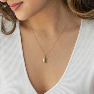 Pu&ograve; includere: Una collana con pendente a medaglione ovale color oro. Il pendente &egrave; appeso a una catena sottile. La collana &egrave; indossata sopra un top bianco a coste.