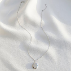 Pu&ograve; includere: Una collana d'argento con un ciondolo rotondo. Il ciondolo &egrave; liscio e riflettente, appeso a una catena delicata. La collana &egrave; presentata su un tessuto bianco strutturato, creando un'estetica minimalista. Il gioiello &egrave; semplice ed elegante.