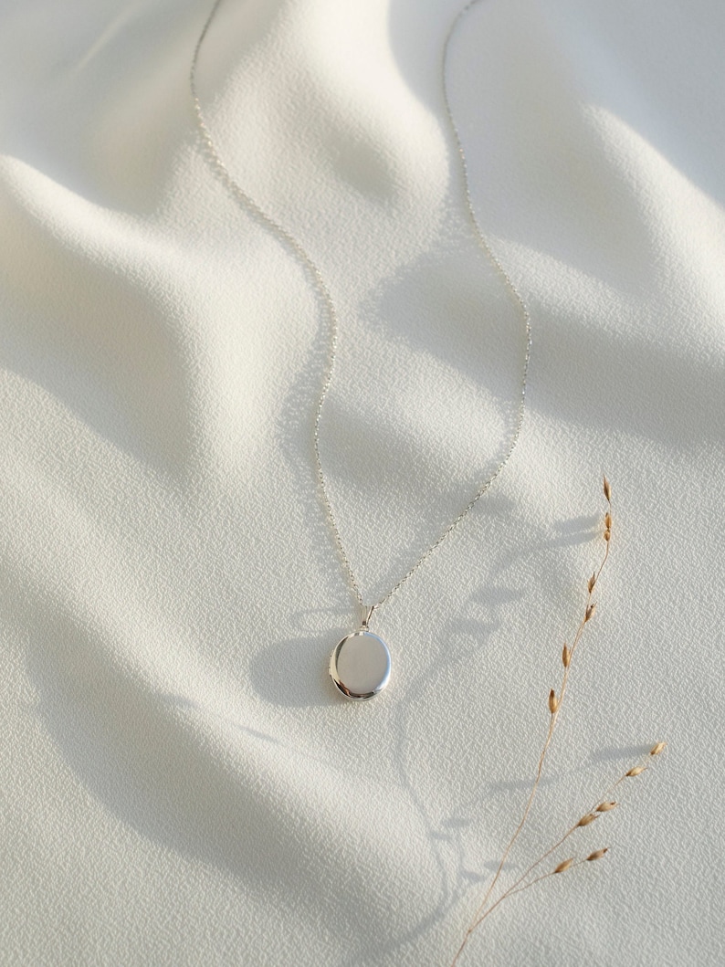 Pu&ograve; includere: Una collana con un medaglione ovale argentato su uno sfondo di tessuto bianco. Il medaglione liscio e riflettente &egrave; sospeso a una delicata catena. Un filo d'erba secca aggiunge un tocco naturale. La collana &egrave; un gioiello.