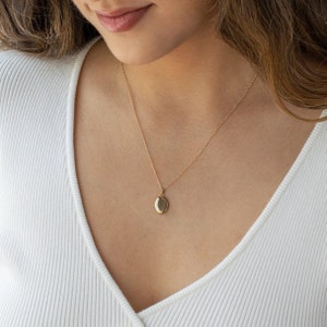 Pu&ograve; includere: Collana con pendente a medaglione ovale dorato. Il pendente &egrave; appeso a una catena sottile. La persona indossa una maglia bianca a maniche lunghe a coste.