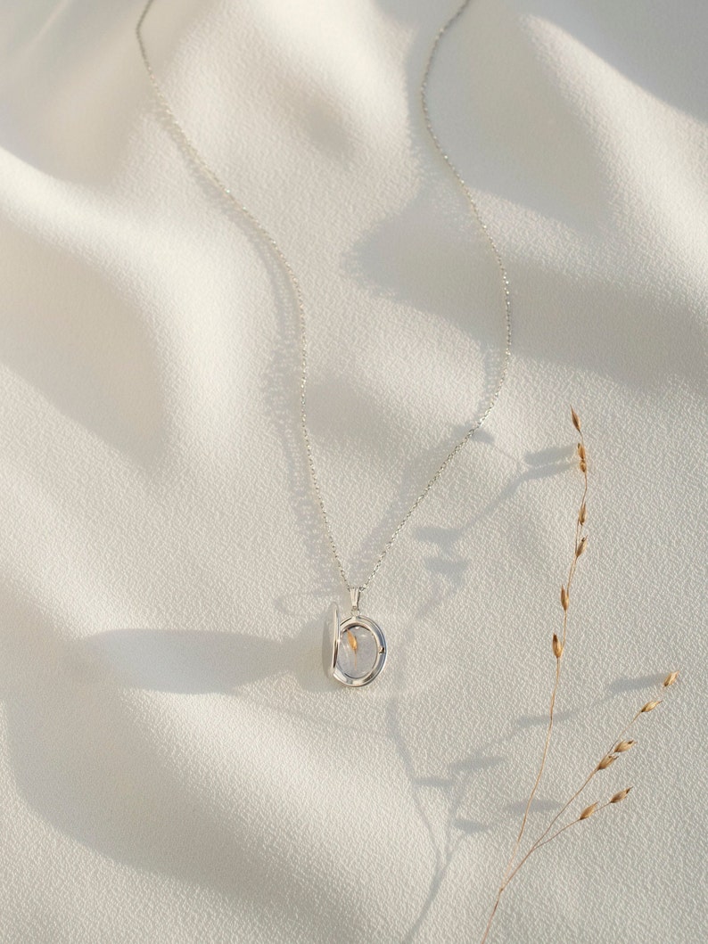Pu&ograve; includere: Una collana d'argento con un piccolo ciondolo rotondo. Il ciondolo ha una parte anteriore trasparente, che rivela un fiore secco all'interno. La collana &egrave; appoggiata su un tessuto bianco strutturato, con un filo d'erba secca sullo sfondo.