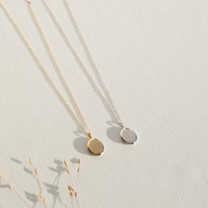 Pu&ograve; includere: Due delicate collane con pendenti ovali. Un pendente &egrave; dorato, l'altro argentato. Entrambe le collane hanno catene sottili e sono esposte su una superficie bianca, con ombre. Le collane sono eleganti e minimaliste.