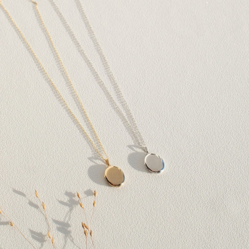 Pu&ograve; includere: Due delicate collane con pendenti ovali. Un pendente &egrave; dorato, l'altro argentato. Entrambe le collane hanno catene sottili e sono esposte su una superficie bianca, con ombre. Le collane sono eleganti e minimaliste.