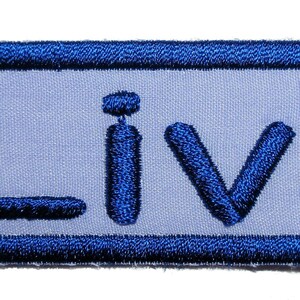 May include: A rectangular embroidered patch featuring a light blue background with a dark blue border. The word Livi is stitched in dark blue thread across the centre of the patch, with the letters L, i, v, and i clearly visible in a bold font.