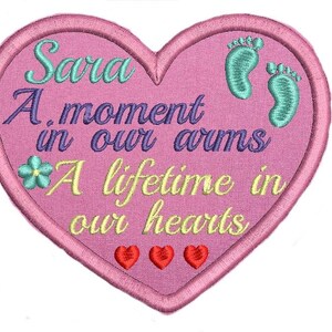 May include: A pink heart-shaped embroidered patch with the text "Sara A moment in our arms A lifetime in our hearts" and two green footprints. There are also three small red hearts at the bottom of the patch.