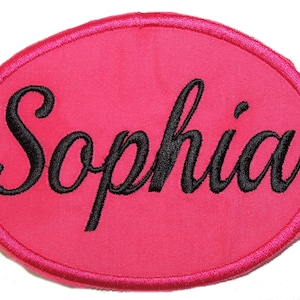 May include: An oval-shaped patch with a vibrant hot pink fabric background and a matching pink stitched border. The name Sophia is embroidered in the centre in a black cursive script font, featuring thick, bold lettering that stands out clearly.
