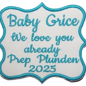 May include: A white embroidered patch with a teal border and teal text. The text reads "Baby Grice, We love you already, Prep Plunden 2025". The patch has a decorative, scalloped edge.