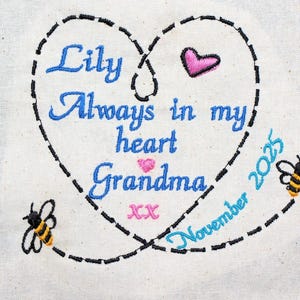 May include: Embroidered heart design with the words "Lily Always in my heart Grandma" in blue script. A pink heart and two bees are also embroidered. The date "November 2025" is included. The design is on a cream-coloured fabric.