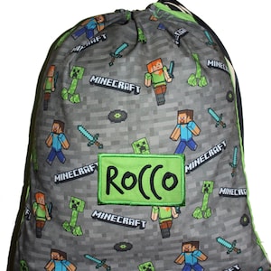 May include: A grey drawstring bag with a Minecraft design. The bag is patterned with pixelated characters, swords, and the word "Minecraft." A green patch with the name "Rocco" is sewn on the front. The bag has a green trim.