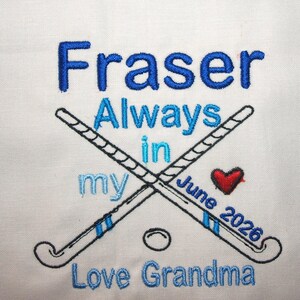 May include: A white fabric surface featuring blue machine embroidery that reads Fraser Always in my June 2026 Love Grandma. The design includes two crossed field hockey sticks in black and blue, a small red heart, and a circular hockey ball outline.
