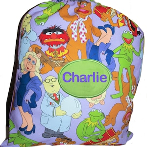 May include: A lavender drawstring bag with a repeating pattern of Muppet characters. The bag features a green oval with the name "Charlie" embroidered in purple. The bag is designed for carrying items.