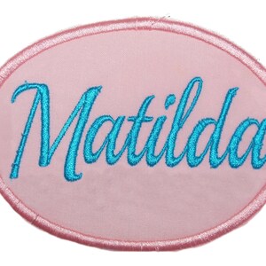 May include: Pink oval-shaped fabric patch with the name Matilda embroidered in blue cursive lettering.