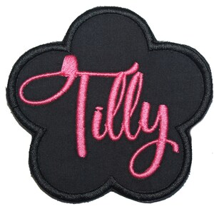 May include: Black, flower-shaped embroidered patch with the name "Tilly" in pink script. The patch has a black border and is set against a white background. This patch is suitable for clothing or accessories.