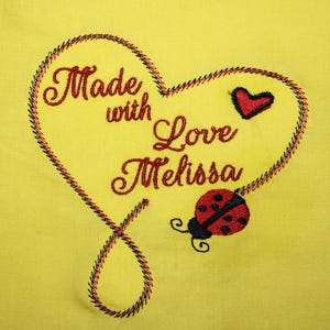 May include: Yellow fabric featuring an embroidered heart design. The design includes the words "Made with Love Melissa" in red script, a small red heart, and a ladybug. The heart is outlined with a red and black stitch.