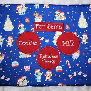 May include: Blue fabric placemat with a Christmas theme featuring cartoon characters and the words "For Santa", "Cookies", "Milk", and "Reindeer Treats".