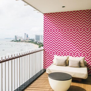 Puede incluir: Balcón exterior con vistas a una ciudad y al océano. El espacio cuenta con un sofá blanco con cojines beige, una mesa blanca y negra, y una pared con un patrón de zigzag rosa y blanco. El suelo es de madera.