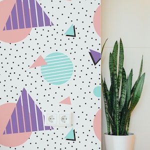 Op de afbeelding: Een muur met een herhalend patroon van geometrische vormen in pastelkleuren en zwarte stippen. Het ontwerp bevat roze cirkels, paarse driehoeken en turquoise vormen. Een plant in een pot met lange groene bladeren staat naast de muur.