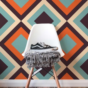 Op de afbeelding: Een witte stoel met houten poten en een deken met franjes houdt een paar zwart-witte sneakers vast. De achtergrond heeft een geometrisch patroon in teal, oranje, beige en donkerpaars.