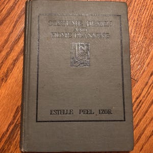 May include: A hardcover book titled Costume Design and Home Planning by Estelle Peel Izor. The book has a muted olive-green fabric cover with dark green lettering and a small rectangular illustration centered on the front cover.