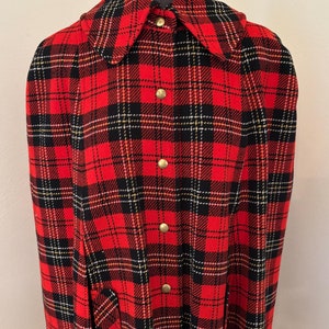 May include: A red and black plaid cape with gold accents and a button closure. The cape has a large collar and two pockets.