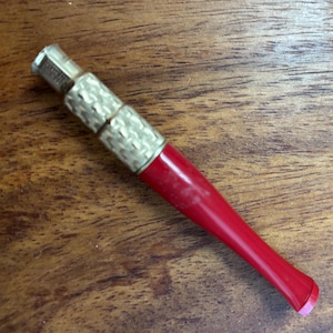 May include: A vintage tobacco pipe with a red stem and a decorative gold-colored metal band. The pipe is resting on a wooden surface, showcasing its classic design and craftsmanship. The pipe's stem is a vibrant red, contrasting with the metallic accents.