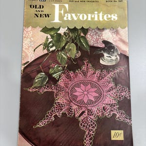 May include: Vintage crochet pattern book titled "Old and New Favorites" with a pink doily on a dark brown table. The cover features the text "Favorites" in large white letters. A small plant and a glass figurine are also on the table.