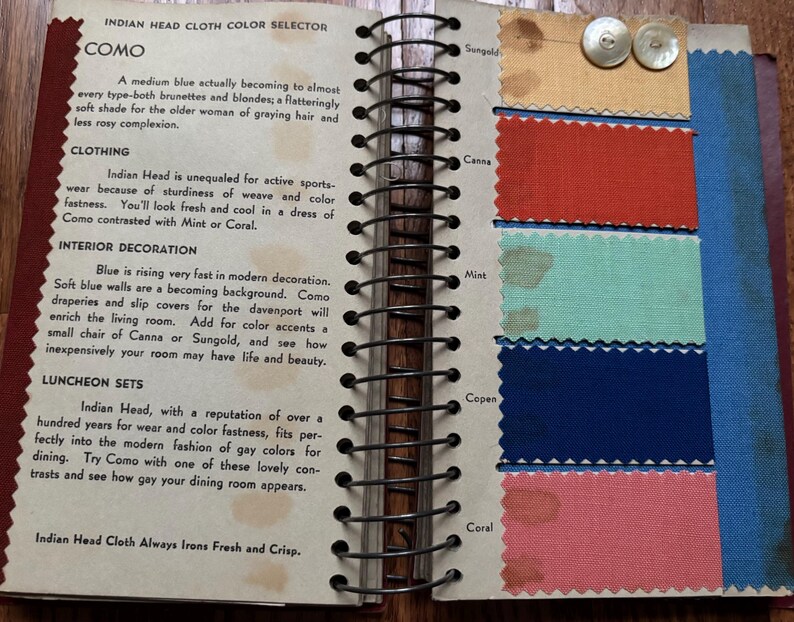 May include: A page from a fabric color selector book showing swatches of different colors of Indian Head cloth. The colors are: Sungold, Canna, Mint, Copen, and Coral. The text on the page reads: "Indian Head Cloth Color Selector, Como, A medium blue actually becoming to almost every type-both brunettes and blondes, a flatteringly soft shade for the older woman of graying hair and less rosy complexion."