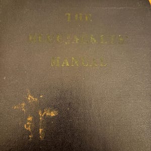 May include: A dark, textured, rectangular book cover with the gold-colored text THE BLUEJACKETS MANUAL printed on the front. The cover features a worn, distressed appearance with visible gold-colored scuff marks on the lower left corner.