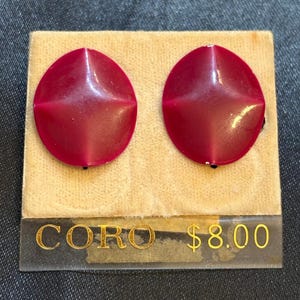 May include: Pair of vintage Coro clip-on earrings. The earrings are a deep red color, round with a geometric design. They are displayed on a beige velvet card with the Coro brand name visible. The price tag reads $8.00.