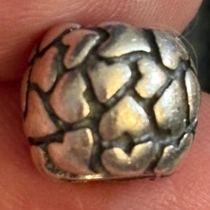 May include: A silver-toned bead with a textured surface. The bead is covered in a pattern of heart shapes. The bead is held between two fingers.