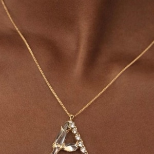 May include: A gold-colored chain necklace with a pendant in the shape of the letter "A". The pendant is embellished with clear crystals. The necklace is displayed against a brown background.