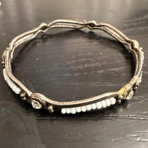 May include: Silver-toned bangle bracelet with a wavy design. The bracelet features small white beads and clear, round gemstones. The metal has a slightly tarnished appearance, giving it a vintage look. The bracelet is circular and appears to be made of metal.