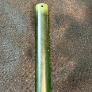 May include: A polished, rectangular jade pendant with a hole at the top for stringing. The pendant is a deep green color with lighter green hues throughout. The smooth surface reflects light, highlighting its natural variations.