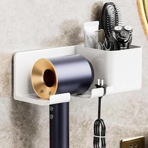 May include: A white wall-mounted organizer for bathroom accessories. It features a holder for a hair dryer with a gold nozzle and a separate compartment for grooming tools, including a brush, comb, and electric shaver.