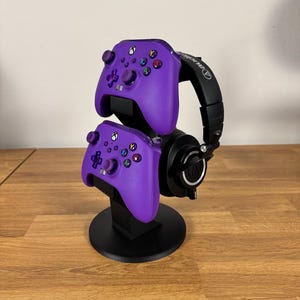 以下が含まれることがあります： 2つの紫色のコントローラーと黒いヘッドフォンを保持する黒いゲームコントローラーのスタンド。スタンドは正方形のベースを持つ円形で、コントローラーは積み重ねられています。ヘッドフォンには白いロゴが付いています。