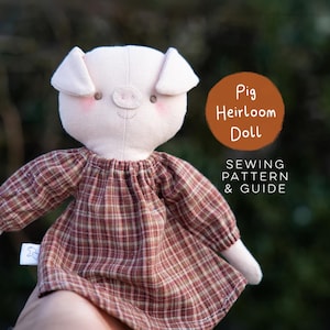 May include: A light pink fabric pig doll wearing a brown and tan plaid dress with long sleeves. The doll has embroidered eyes and pink cheeks. Text on the image reads: Pig Heirloom Doll SEWING PATTERN & GUIDE. The doll has a small white side tag.