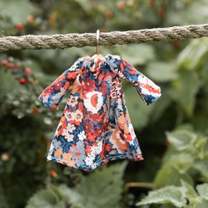 Puede incluir: Un vestido en miniatura con un vibrante estampado floral en tonos rojos, azules, blancos y naranjas. La prenda tiene mangas largas, un escote fruncido y est&aacute; colgada de una peque&ntilde;a percha de metal en una cuerda gruesa y texturizada.
