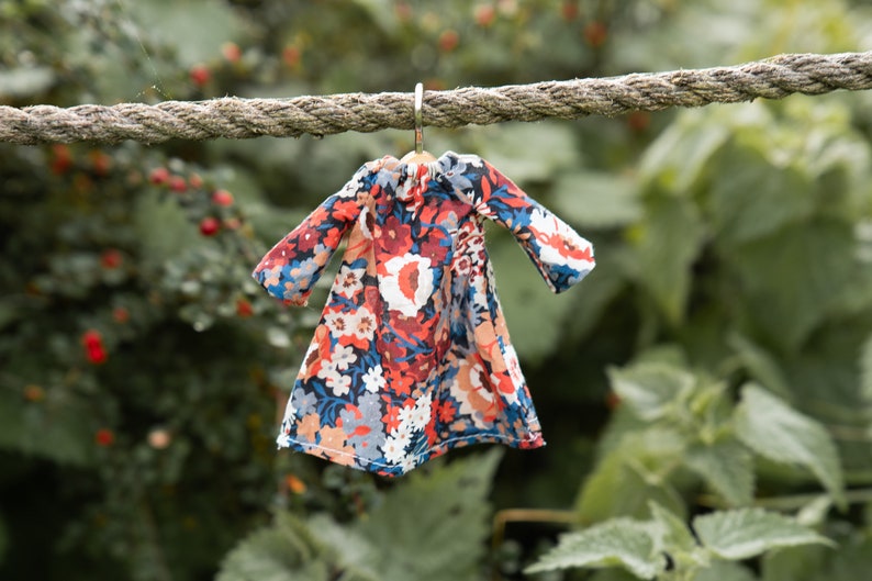 Puede incluir: Un vestido en miniatura con un vibrante estampado floral en tonos rojos, azules, blancos y naranjas. La prenda tiene mangas largas, un escote fruncido y est&aacute; colgada de una peque&ntilde;a percha de metal en una cuerda gruesa y texturizada.