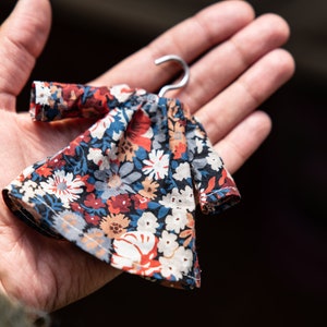 Puede incluir: Un vestido en miniatura con fondo azul marino oscuro y un denso estampado floral en tonos rojo, blanco, azul claro y beige. La prenda incluye mangas largas y est&aacute; suspendida de una peque&ntilde;a percha de metal de color plateado.