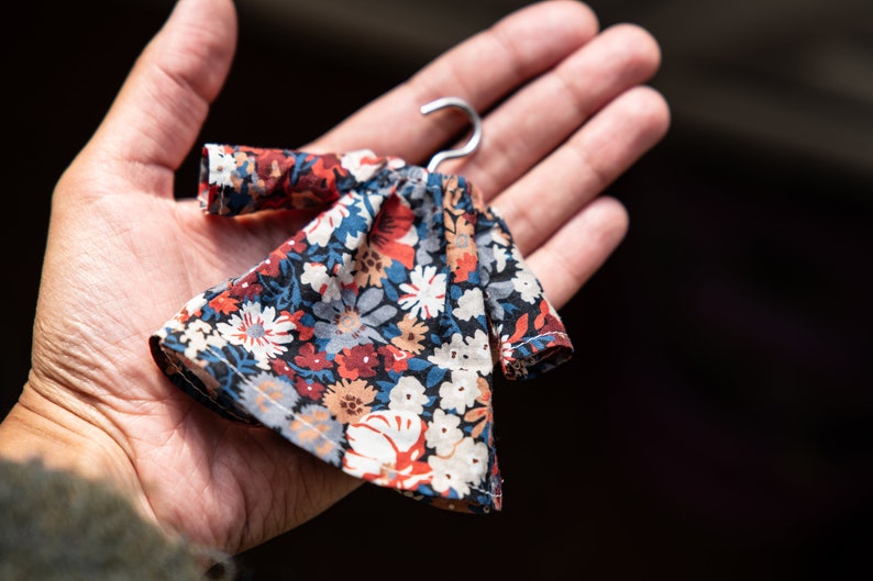 Puede incluir: Un vestido en miniatura con fondo azul marino oscuro y un denso estampado floral en tonos rojo, blanco, azul claro y beige. La prenda incluye mangas largas y est&aacute; suspendida de una peque&ntilde;a percha de metal de color plateado.