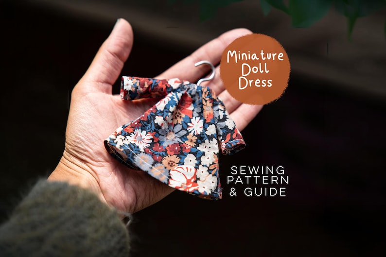 Puede incluir: Un vestido de mu&ntilde;eca en miniatura con un estampado floral oscuro de flores rojas, blancas y azules sobre un fondo negro, colgado de una peque&ntilde;a percha de metal. El texto en la imagen dice Miniature Doll Dress SEWING PATTERN & GUIDE.