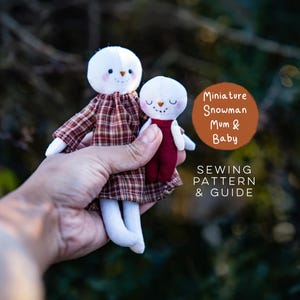 May include: Two fabric snowman dolls, one larger and one smaller, featuring white heads with embroidered faces and rosy cheeks. The larger doll wears a brown plaid dress, and the smaller doll wears a solid maroon outfit. Text reads Miniature Snowman Mum & Baby SEWING PATTERN & GUIDE.