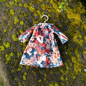 Puede incluir: Un vestido en miniatura sobre una peque&ntilde;a percha de metal con un denso estampado floral en tonos azul oscuro, rojo intenso, melocot&oacute;n y blanco. La prenda tiene mangas largas, un cuello fruncido y una falda acampanada, dise&ntilde;ada para una mu&ntilde;eca.
