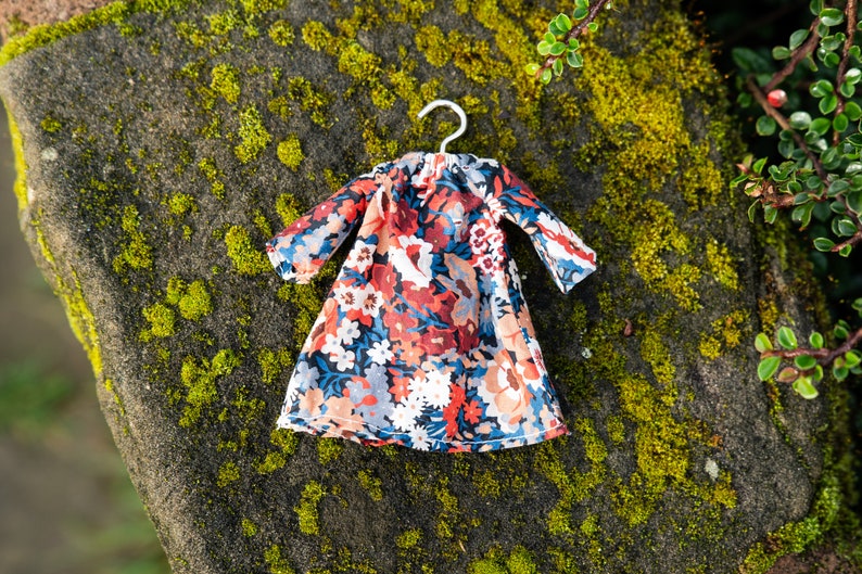 Puede incluir: Un vestido en miniatura sobre una peque&ntilde;a percha de metal con un denso estampado floral en tonos azul oscuro, rojo intenso, melocot&oacute;n y blanco. La prenda tiene mangas largas, un cuello fruncido y una falda acampanada, dise&ntilde;ada para una mu&ntilde;eca.