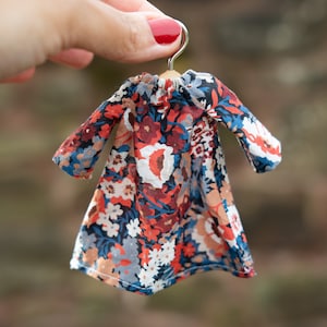 Puede incluir: Un vestido en miniatura de manga larga con un denso estampado floral en tonos azul oscuro, rojo intenso, blanco y naranja claro. La prenda cuelga de una peque&ntilde;a percha de madera con un gancho de metal plateado y presenta un cuello fruncido.