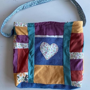 May include: A patchwork tote bag with a blue denim base and a heart-shaped patch. The bag has a variety of colors and patterns, including red, orange, yellow, green, purple, and white. The bag has a long strap.