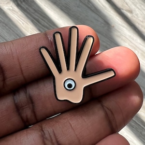 May include: A black and brown enamel pin shaped like a hand with an eye in the palm.