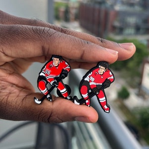 May include: Two red and black enamel hockey player pins. The players are in a skating position with a hockey stick. The number 98 is on the front of each player's jersey.