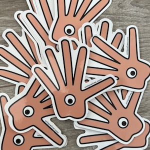 May include: A collection of stickers depicting a cartoon hand with an eye. The hand is peach-coloured with black outlines and a white border. The text "unique3ree.com" is printed on the stickers.